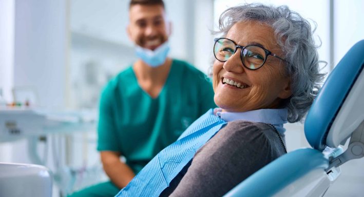Satisfied senior woman at dentist’s office looking at camera. Happy senior woman at dental clinic looking at camera. Her dentist is in the background.